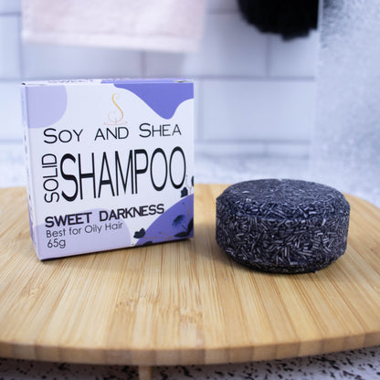 A white box with purple abstract pattern and the text SOLID SHAMPOO SWEET DARKNESS stands upright on a wooden tray with a blurred bathroom background.  Next to the box sits a round bar of solid shampoo.  The bar is dark purple with small white lines throughout.