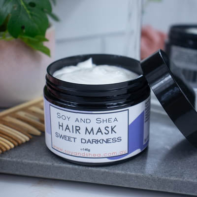 A black jar with the lid resting on its side revealing a white cream inside, sits on a dark grey tile surface against a blurred background of a bathroom counter top. The jar features a white label with purple abstract design with text HAIR MASK SWEET DARKNESS.