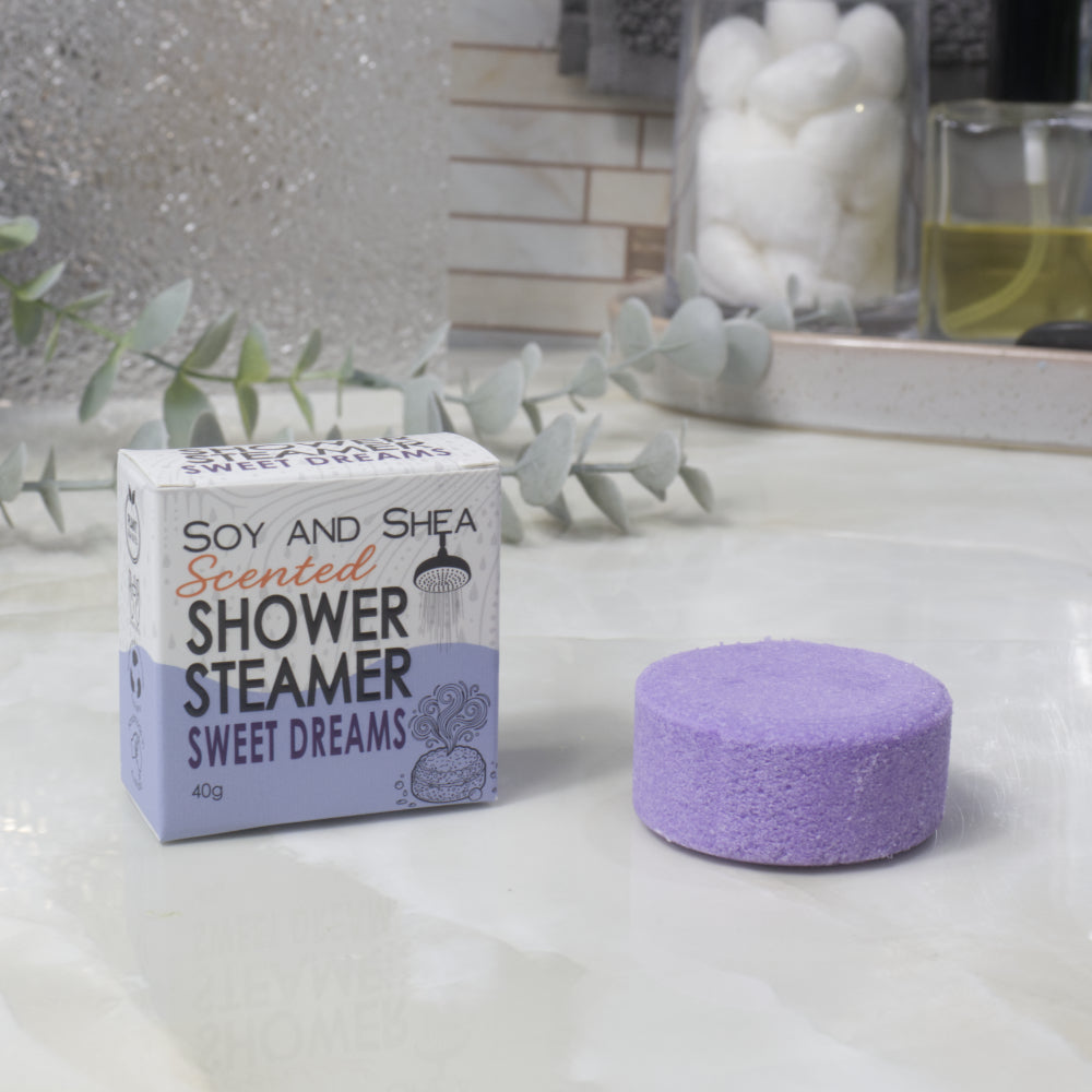 A boxed scented shower steamer and the steamer itself on a bathroom counter. The box reads 'Soy and Shea Scented Shower Steamer Sweet Dreams', and the round purple steamer sits beside it.