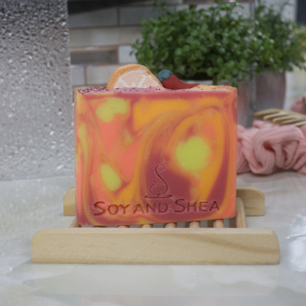 The Sweet Orange Chilli Pepper Soap Bar by Soy and Shea, featuring orange, yellow, and red swirls, rests on a wooden soap dish on a bathroom counter.  A plant and scrunchie can be seen in the background against a tiled wall. 
