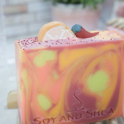 A close up photo of a soap bar with red, orange and yellow swirls.  The picture focuses on the Soap embellishments stuck into the top of the soap featuring a slice of orange and small chilli pepper made with soap