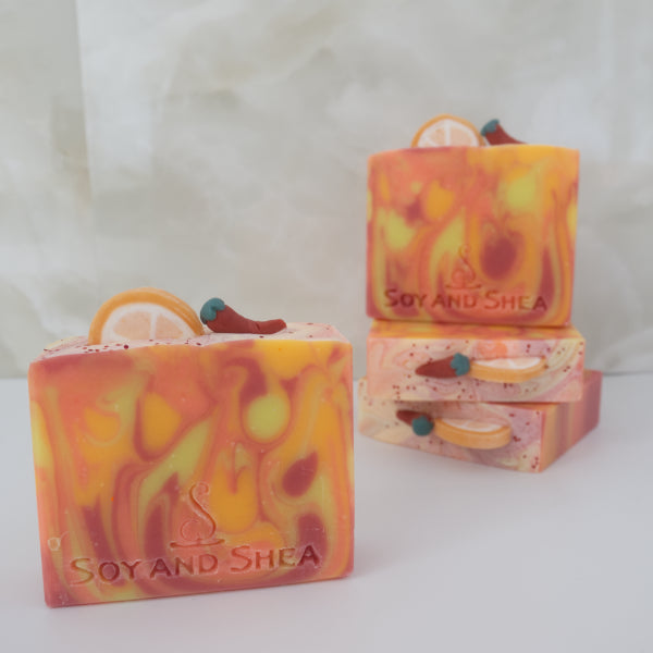 A collection of four soaps sit on marble tiles.  Each soaps shows a unique design of red, orange and yellow swirls.    Each soap features soap embellishments that are of a round orange slice and small chilli