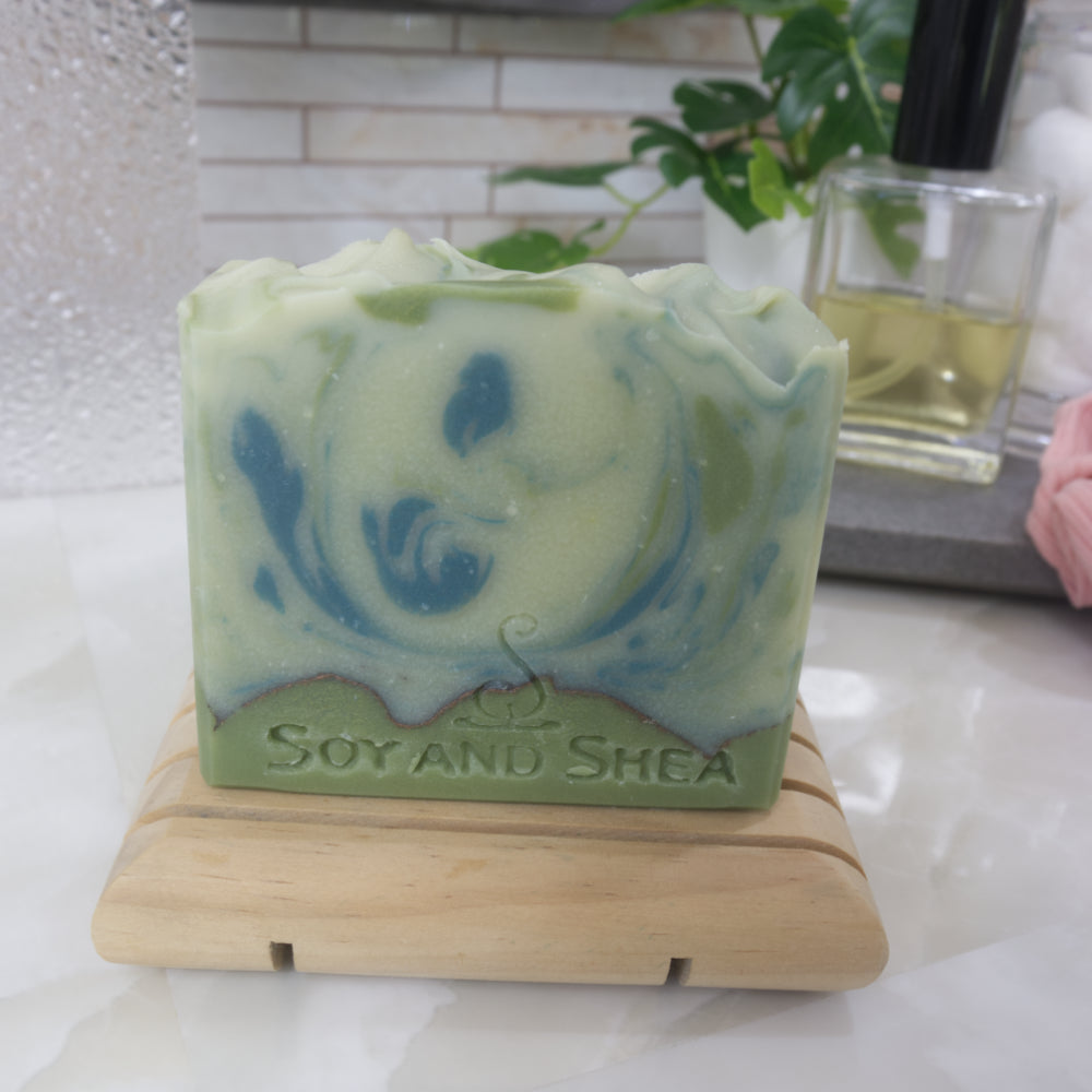 Pale green soap bar with dark green swirls on a wooden soap rack on a bathroom counter with blurred background
