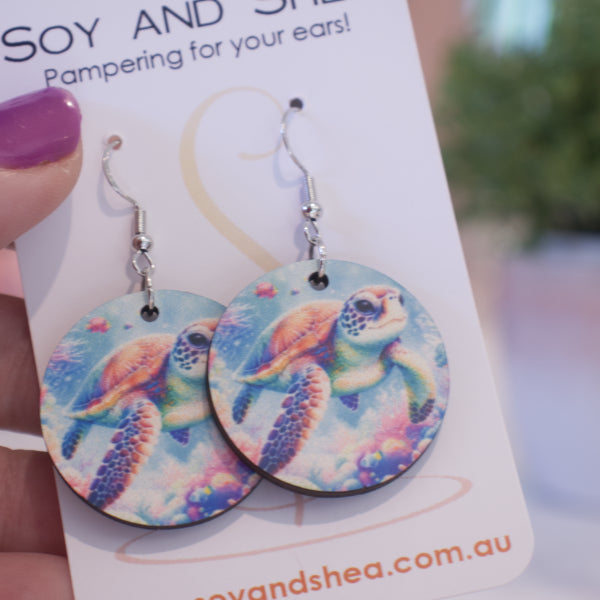 A hand holds a white card branded with SOY AND SHEA with a pair of earrings hanging on hooks.  The earrings are round with a watercolour image of a turtle swimming over a reef