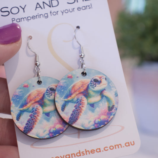 A hand holds a white card branded with SOY AND SHEA with a pair of earrings hanging on hooks.  The earrings are round with a watercolour image of a turtle swimming over a reef