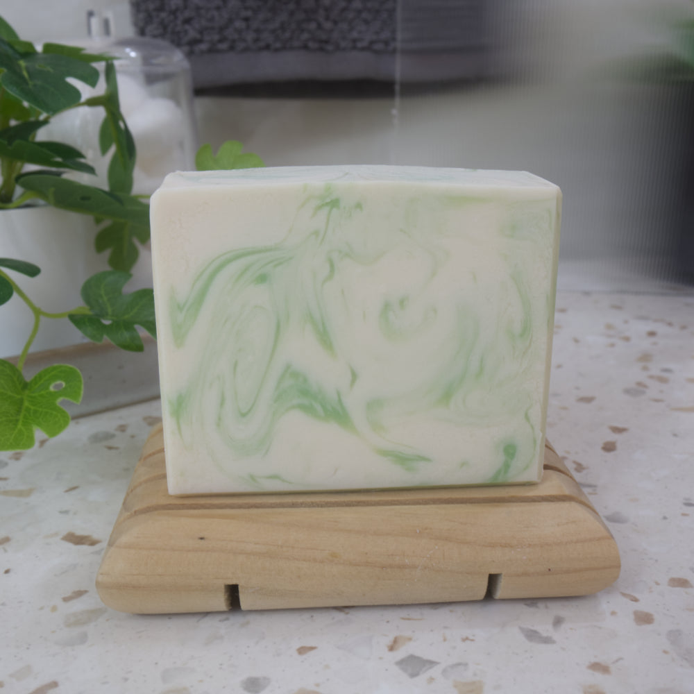 A white soap bar with green swirls stands on a wooden soap dish on a batrhroom counter along with a green plant.
