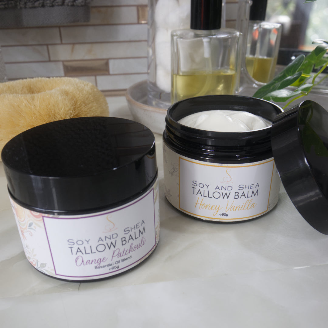 Two black jars of Tallow Balm with decorative labels are sitting on a bathroom counter with a bottle of purfume and body brush.  The jar towards the front left has a black lid and a label identifying it as Orange Patchouli.  To the back right of this jar is another which has the lid removed and resting on the side of the jar revealing a white balm.  This is labelled Honey Vanilla
