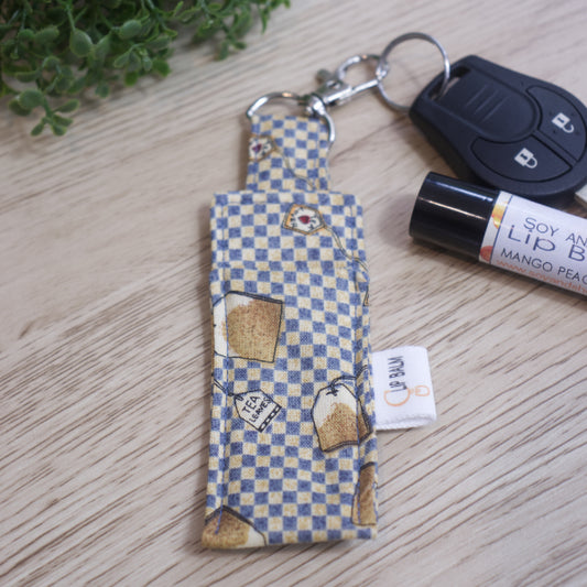 A Lip Balm holder lies on an wooden surface with its lobster claw hook attached to a car key.  A lip balm and greener can be seen in the image.  The Clip Balm has a blue and cream checkered background covered with illustrations of tea bags.