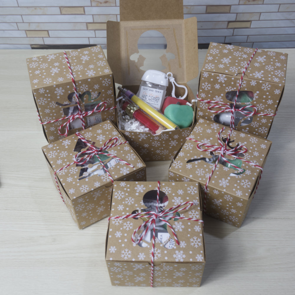 Five small brown kraft gift boxes with white snowflake patterns and tied with red twine are arranged on a light wood surface. A sixth box is open in the center, showing small assorted items inside including two apple shaped soaps, a pencil shaped lip oil, a pen and a small bottle of hand cleanser