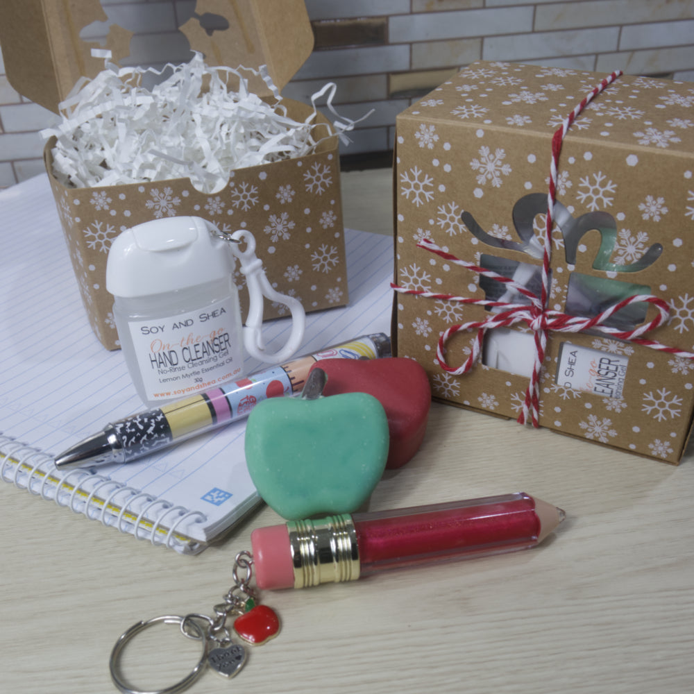 A curated gift set featuring a Soy and Shea hand cleaner, a red lip oil keychain in the shape of a pencil, a green and red apple-shaped soap, a decorative pen, and wrapped gift boxes with white paper shred, arranged on a light wood tabletop.