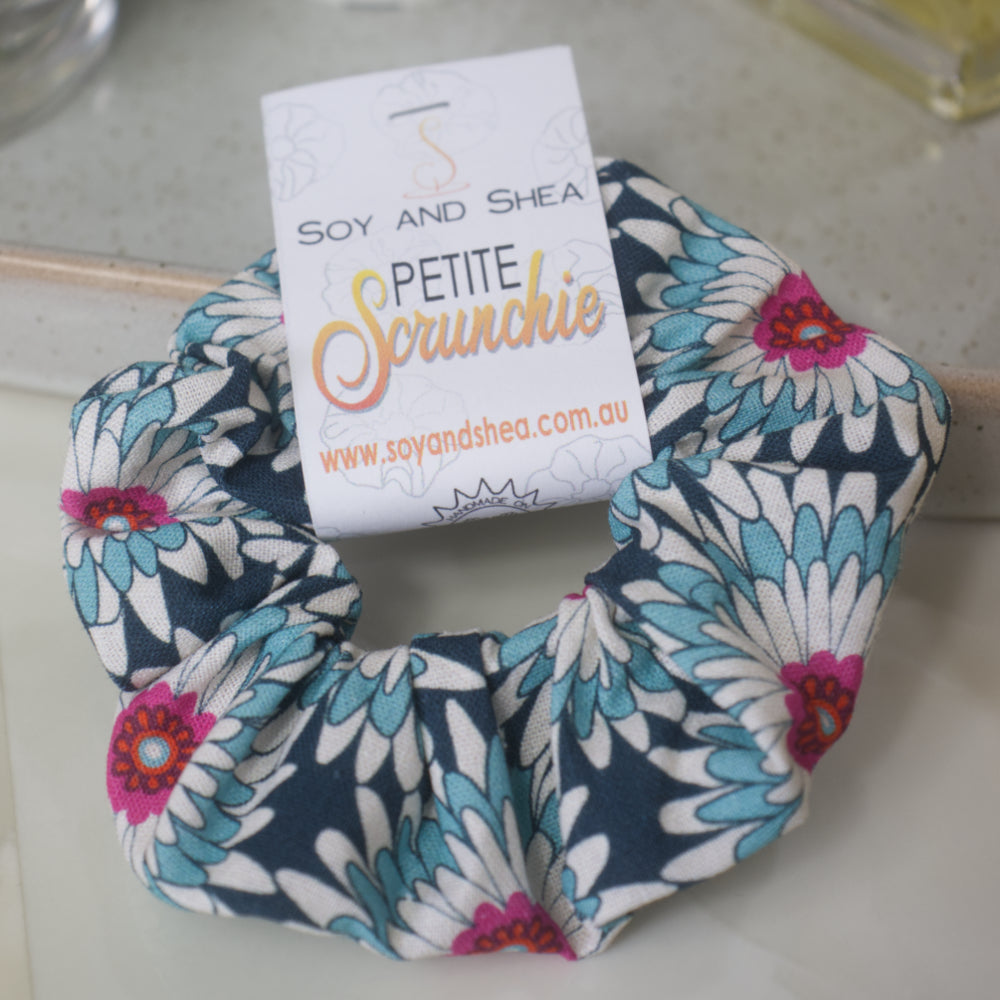 A small scrunchie with product label identifying it as Petite, lays on a marble surface resting against a stone tray.  The scrunchie features gerbera like flowers in shades of dark blue, white and teal with bright pink centers.