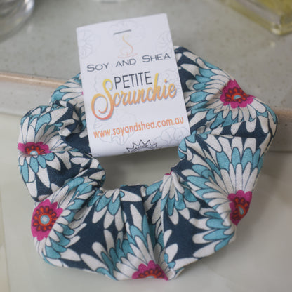 A small scrunchie with product label identifying it as Petite, lays on a marble surface resting against a stone tray.  The scrunchie features gerbera like flowers in shades of dark blue, white and teal with bright pink centers.