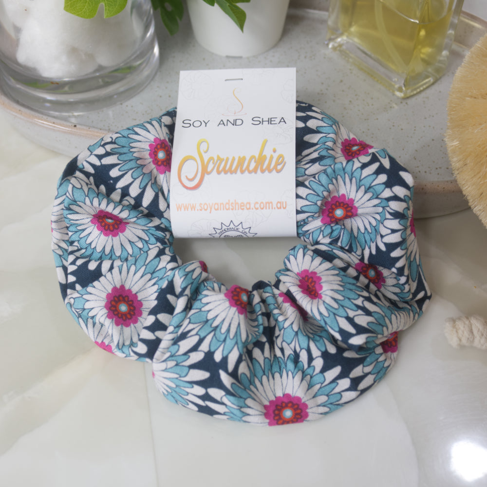A scrunchie lies on a bathroom counter top resting against a stone tray with perfume bottle and jar.  The scrunchie features gerbera like flowers in shades of dark blue, white and teal with bright pink centers.