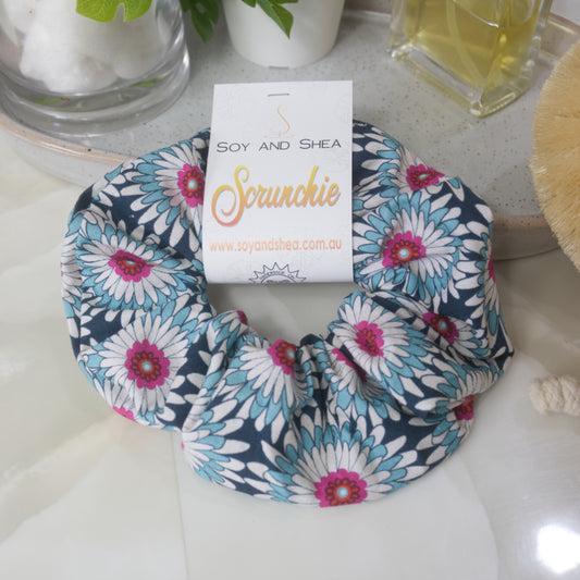 A scrunchie lies on a bathroom counter top resting against a stone tray with perfume bottle and jar.  The scrunchie features gerbera like flowers in shades of dark blue, white and teal with bright pink centers.