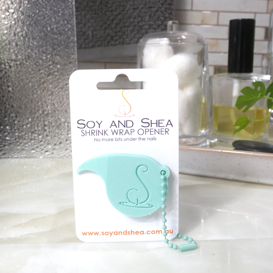 A Teal Shrinkwrap opener with matching ball and chain keyring is attached to a white card with product details.  The card is standing upright on a bathroom countertop with a bottle of perfume, a jar of cotton balls and towel in the background