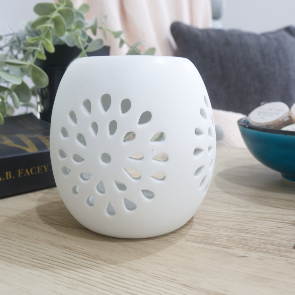 An unlit matte white ceramic fragrance warmer sits on a light wooden table, turned to fully display the intricate, circular starburst pattern of teardrop-shaped cutouts on its side. To the left is a black book and green foliage, and to the right is the edge of a blue ceramic bowl with wrapped wax melts. The background features a softly blurred dark grey cushion and a pink textured throw blanket.