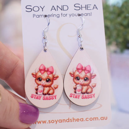 A hand holds a white card branded with SOY AND SHEA with a pair of earrings hanging on hooks.  The earrings are teardrop shaped  with a pale pink background.  An illustration of a baby highland cow with pink bow on head drinking from a white cup with heart and straw sits above the words STAY SASSY