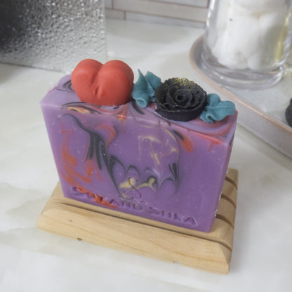 A soap bar stands on a wooden soap rack on a bathroom counter surrouned by perfume, a jar with cotton balls and a plant.  The soap is bright purple with swirls of red, gold and back.  The image focuses on the top of the soap which has embellishments made of soap in the shape of a red heart and a black rose with green leaves.
