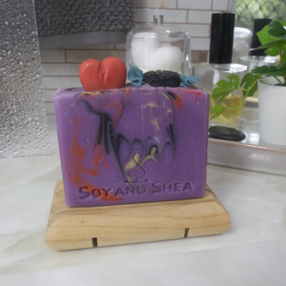A soap bar stands on a wooden soap rack on a bathroom counter surrouned by perfume, a jar with cotton balls and a plant.  The soap is bright purple with swirls of red, gold and back.  The top of has embellishments made of soap in the shape of a red heart and a black rose with green leaves.