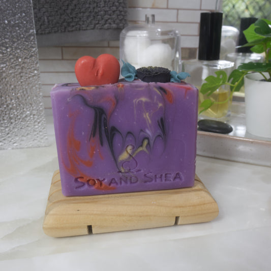 A soap bar stands on a wooden soap rack on a bathroom counter surrouned by perfume, a jar with cotton balls and a plant.  The soap is bright purple with swirls of red, gold and back.  The top of has embellishments made of soap in the shape of a red heart and a black rose with green leaves.