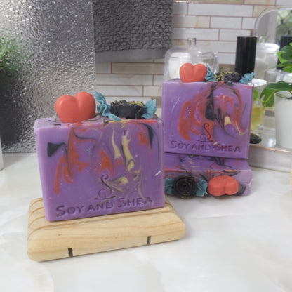 A selection of three soaps displayed on a bathroom bendch along with a bottle of perfume, plant and jar of cotton balls.  One soap sits on a wooden rack to the front left of the image, another two are displayed behind with one laying flat showing the top of the soap with a third standing on top. Each soap is a vivid purple with various swirls of red, black and gold.  Each soap is topped with soap embellishments of a red heart and a black rose with green leaves.