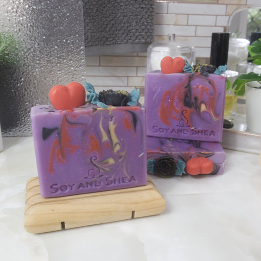 A selection of three soaps displayed on a bathroom bendch along with a bottle of perfume, plant and jar of cotton balls.  One soap sits on a wooden rack to the front left of the image, another two are displayed behind with one laying flat showing the top of the soap with a third standing on top. Each soap is a vivid purple with various swirls of red, black and gold.  Each soap is topped with soap embellishments of a red heart and a black rose with green leaves.