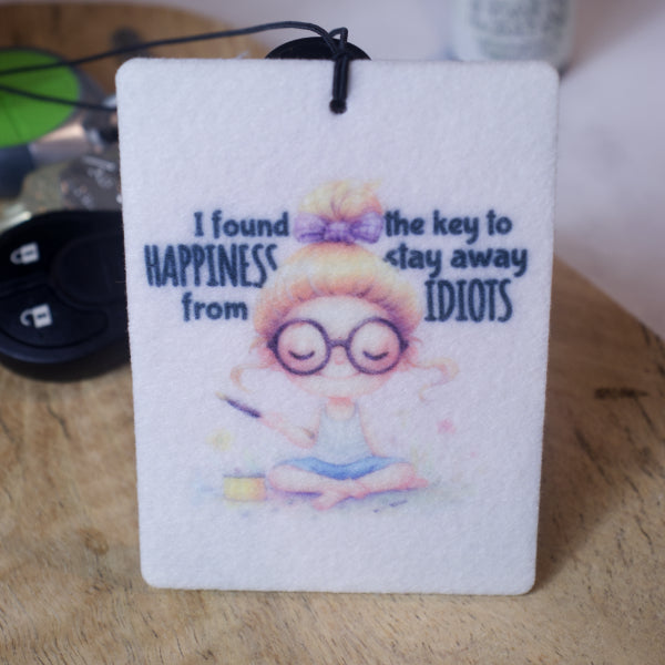 A rectangular car air freshener stands on a wooden surface with a set of car keys in the background.  The Air Freshener  has a cream colour with a watercolour image of a young woman sitting cross legged on a patch of grass holding a stick of incense.  Around her head are the words I FOUND THE KEY TO HAPPINESS, STAY AWAY FROM IDIOTS.