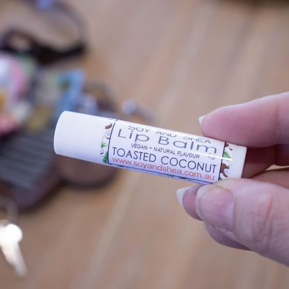 A hand holds a white tube labelled lip balm over a blurred image of an open handbag with contents spilling onto the table.  The lip balms features white label with a random pattern of coconut halves