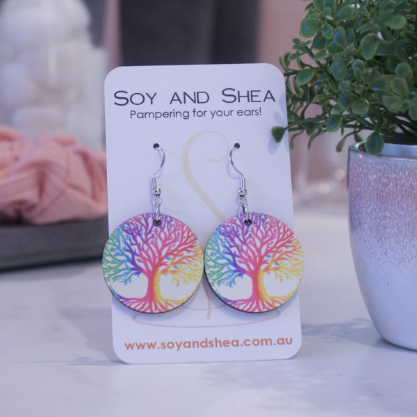 A pair of earrings on silver hooks hang on a white card branded with SOY AND SHEA leaning against a pot with a green leafy plant and a blurred background.  The earrings are round featuring a rainbow colours tree of life stretching across the whole earrings against a white background