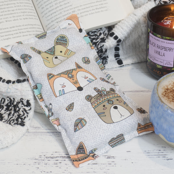 A small fabric heat pack placed on a wooden surface rests against a black and white blanket with open book, along side a burning candle and a filled coffee cup.  The Heat Pack features a collection of small cartoon animal heads including bears and foxes wearing Indian style feathered headbands agains a white and grey background.  Small feathers are scattered between the larger images