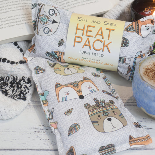 Two Tribal Cubs Mini Heat Pack by Soy and Shea rests near a ceramic coffee cup, a burning candle, and an open book—creating an inviting cosy scene.  The Mini Heat packs feature a white backgroudn with grey speckles.  Scattered through our are drawings of cub like animals including bears, foxes and owls with indian like headresses.  There are feathers scattered around them.