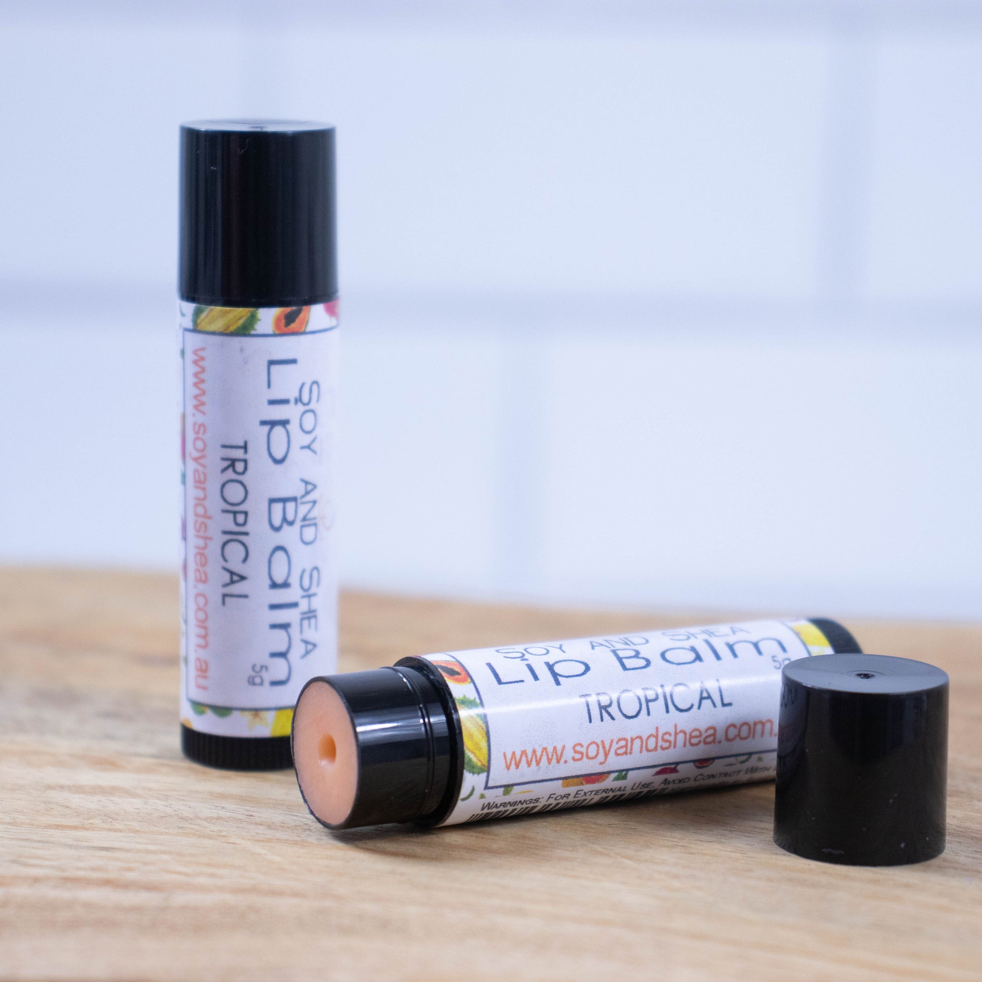 Two black tubes of lip balm are displayed on a wooden surface with blurred tiled background.  One tube stands on its end while the other lays flat in front with the cap removed revealing a orange balm.  The label is white with illustrations of tropical fruits