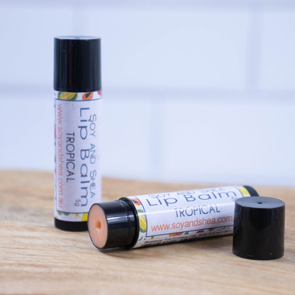 Two black tubes of lip balm are displayed on a wooden surface with blurred tiled background.  One tube stands on its end while the other lays flat in front with the cap removed revealing a orange balm.  The label is white with illustrations of tropical fruits