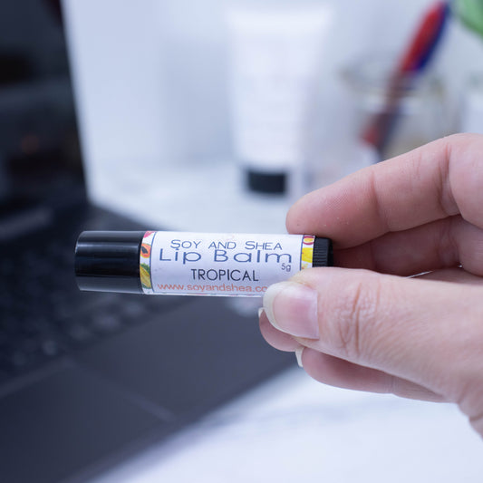 A hand holds a black tube of lip balm against a blurred background with an open laptop computer on a white surface.  The label has text LIP BALM TROPICAL on a white background with a border of tropical fruits in yellow and orange.
