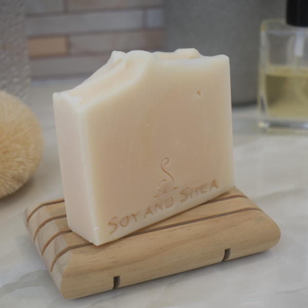 A cream coloured soap bar sits on a wooden soap rack on a bathrooom counter top with tilled background and blurred accessories