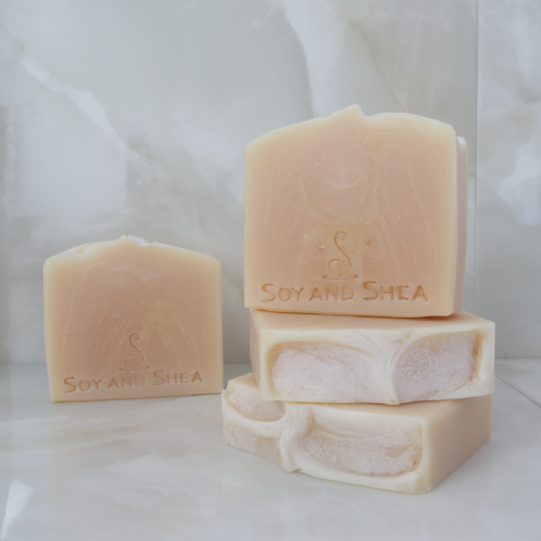 In the foreground, two soaps lay flat on a marble tile surface showing a textured top.  Another soap stands on top with a fourth in the background agains a tile background.  All soaps are plain cream colour with a slightly lighter colour swirl throughout.