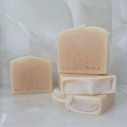 In the foreground, two soaps lay flat on a marble tile surface showing a textured top.  Another soap stands on top with a fourth in the background agains a tile background.  All soaps are plain cream colour with a slightly lighter colour swirl throughout.