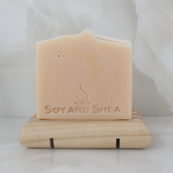 A cream coloured soap bar stamped with SOY AND SHEA sits on a wooden soap dish against a marble tile background.