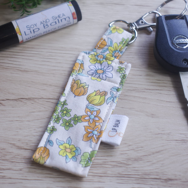A lip balm holder lies on a wooden surface attached to a car key with a "lobster claw" clip.  To the side is a tube of lip balm. The Clip Balm has a cream fabric covered in various flowers including orange and yellow tulips.