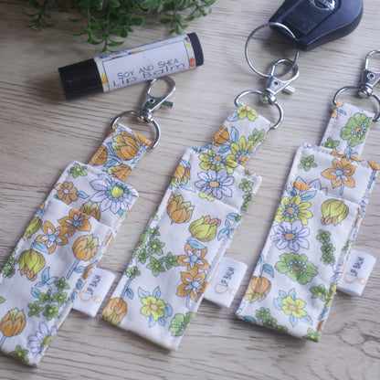A selection of three clip balms showing variations in fabric design, lie on a wooden side with the middle Clip Balm attached to a car key.  A lip balm lays near-by along with some greenery.  The Clip balms have illustrations of yellow, orange, green and white flowers in cluding several orange tulips all on a cream background.  Each Lip Balm Holder has a slight variation in pattern