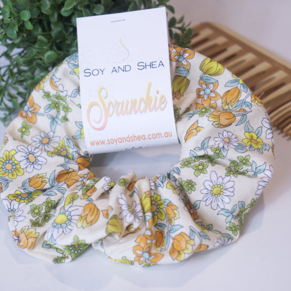 A colorful scrunchie sits on wooden surface resting against a wooden comb with greenery in the background.  A cardboard tag reads SOY AND SHEA SCRUNCHIE.    The scrunchie is a cream fabric with yellow/orange tulips and white daisies randomly placed with small green leaves