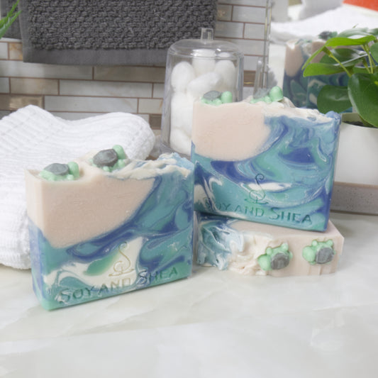 Three Soap bars are displayed on a bathroom countertop along with a towel and greenery.  One soap lays flat on the bench showing the top of the soap with two small turtles on beige soap. On top of this stands another soap and the left front, a third stands on the counter,   The soaps have a ocean like swirl of blue colours and a small patch of beige soap in the top left corner. All feature two small turtles on the beige part.