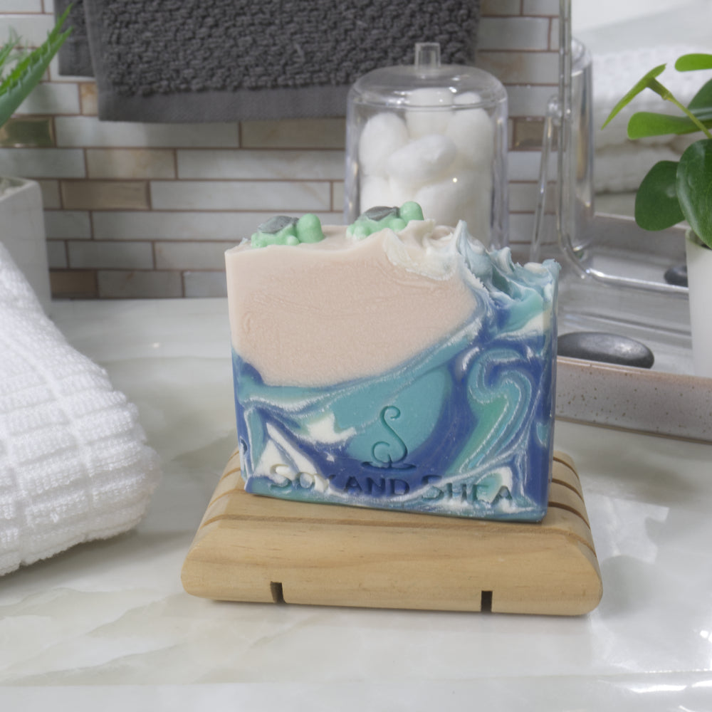 A bar of soap stands on a wooden soap rack on a bathroom counter top.  The soap features a swirling pattern of ocean blues with a beige left corner.  On top of the beige section is two small turtle shapes
