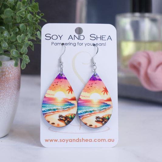 A pair of earrings on silver hooks hang on a white card branded with SOY AND SHEA leaning against a pot with a green leafy plant and a blurred background.  The earrings are teardrop shaped showing a beach at sunet.  Along the sand, a turtle can be seen moving towards the water which has shades of teal moving to purple as it gets closer to the horizon when a bright sun is setting agains a orange/pink sky
