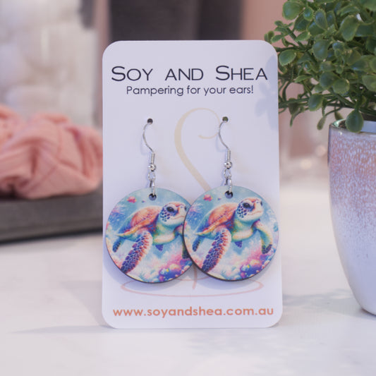 A pair of earrings on silver hooks hang on a white card branded with SOY AND SHEA leaning against a pot with a green leafy plant and a blurred background.  The earrings are round with a watercolour image of a turtle swimming over a ocean floor covered in coral 