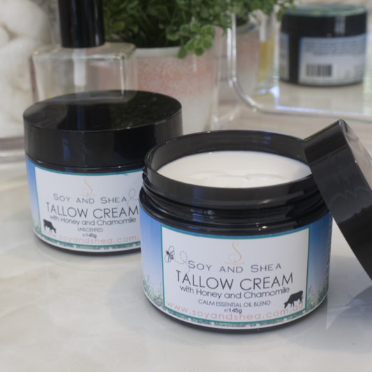 Two black jars of Tallow Cream, one with the lid off showing the white cream inside, and the other with the lid on showing the label that reads 'SOY AND SHEA TALLOW CREAM' sit on a bathroom counter top with perfume bottle, mirror and plant.