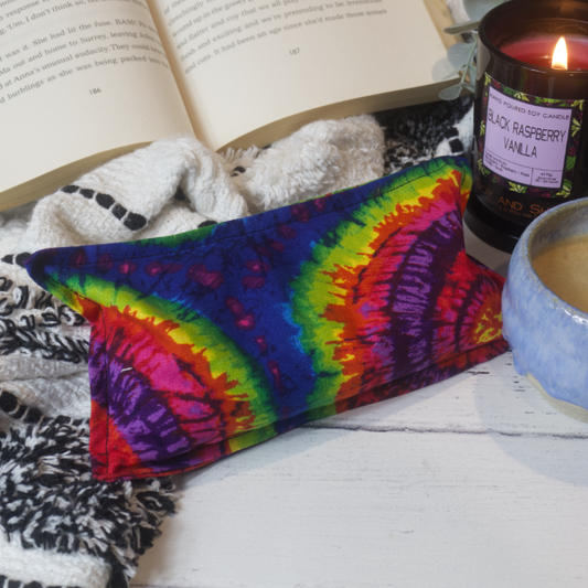 A mini rectangular heat pack rests against a black and white blanket and open book.  Next to it is a burning candle and filled coffee cup evoking a comfy scene.   The heat pack features a tie dyed pattern of concentric circles in rich rainbow colours.