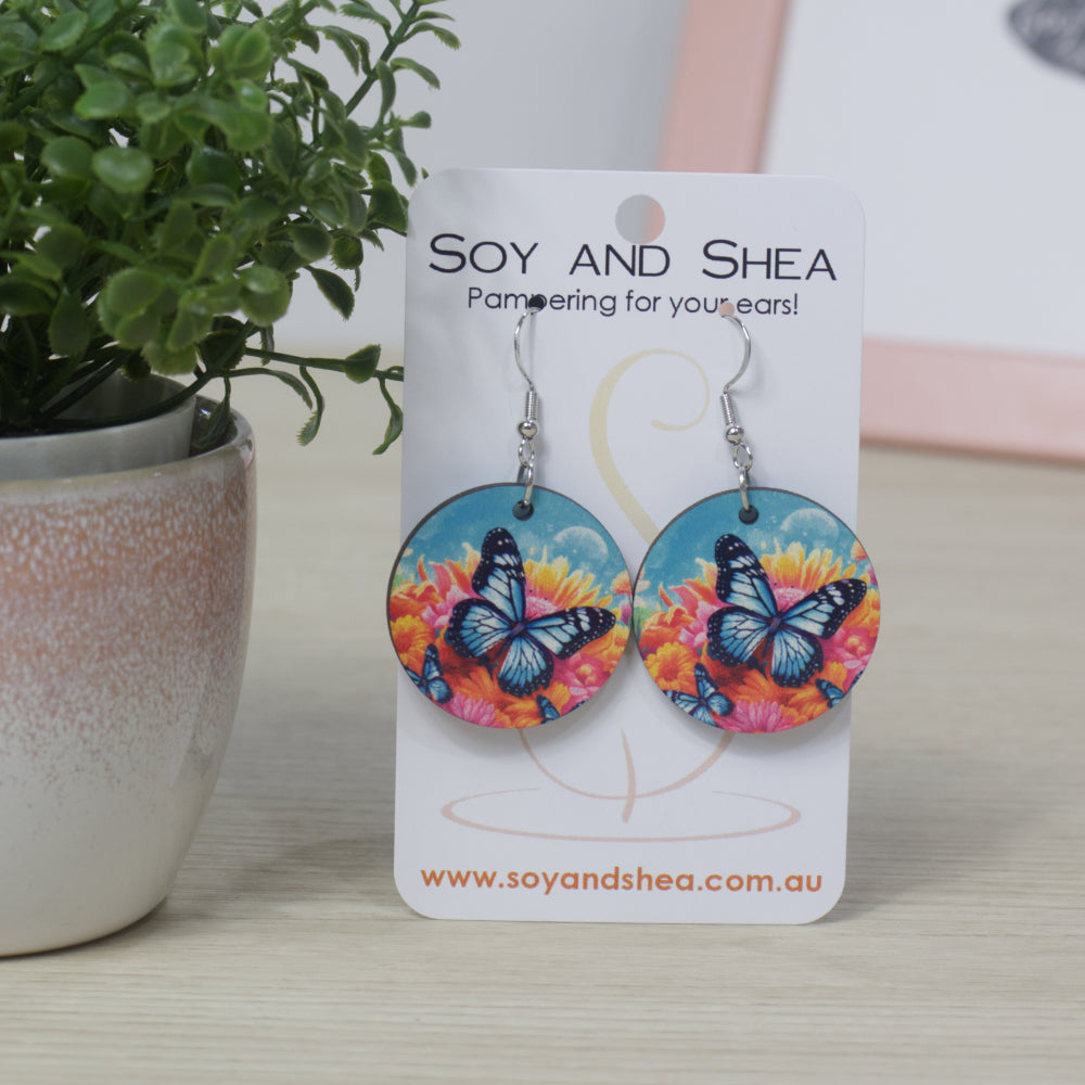 The Ulysses Earrings by Soy and Shea feature round sublimated designs with blue butterflies on colorful flowers, sterling silver hooks for sensitive ears, and come displayed on a branded card. 
