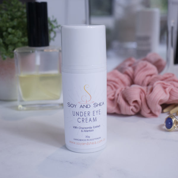 A white bottle with white cap and label reading UNDER EYE CREAM sits on a bathroom counter top surrounded by a pink scrunchie, bottle of perfume and plant with a mirror reflecting the scene