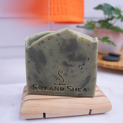 A soap bar sits on a wooden soap dish on bathroom counter top with a plant and towel blurred in the background.  The Soap is dusky green with darker green drops along with brown flecks 